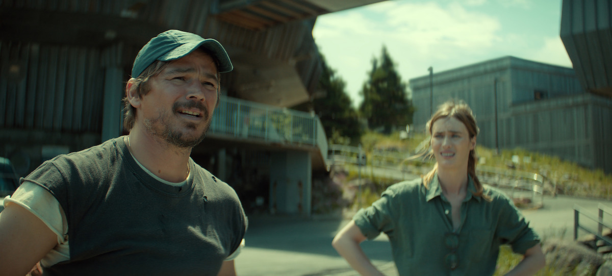 Two people stand outdoors near modern industrial buildings on a sunny day. The man wears a cap and casual clothes, while the woman stands with her hands on her hips, both looking in the same direction.