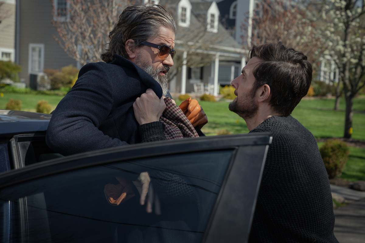 A man gripping another man's coat, confronting him beside a car parked in a suburban neighborhood with houses and trees in the background on a clear day.