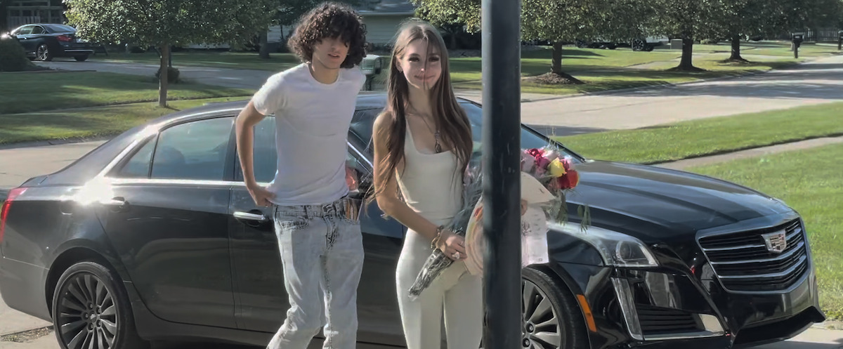 Two teenagers standing by a black Cadillac on a suburban street, with the girl holding flowers and both dressed in light-colored clothes, trees and grass visible in the background.