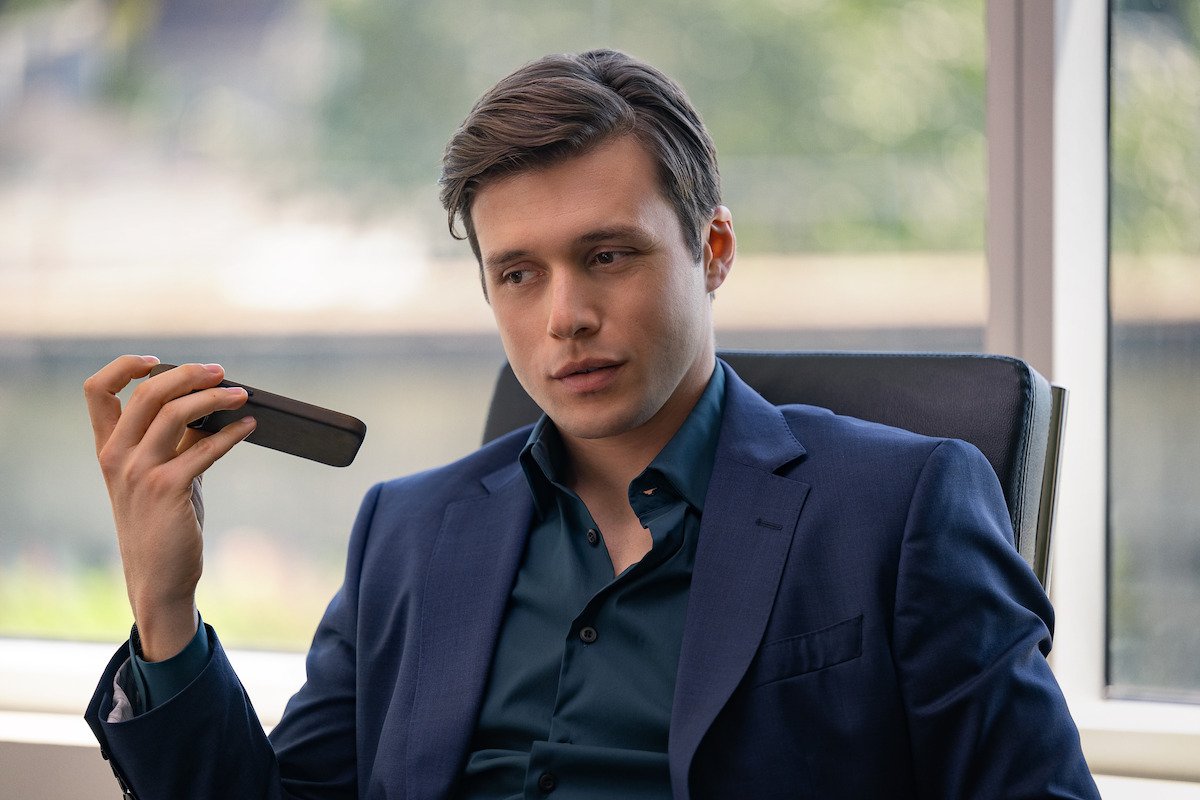 Man in a business suit sitting in an office, holding a phone on speaker with a serious expression, natural light coming in through large window, blurred greenery outside.