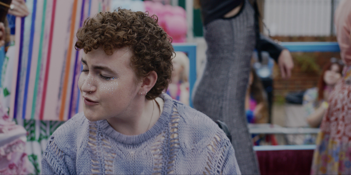 A person with curly hair and glitter on their face wears a textured sweater, sitting at an outdoor gathering with colorful decorations and people, suggesting a festive or celebratory environment.