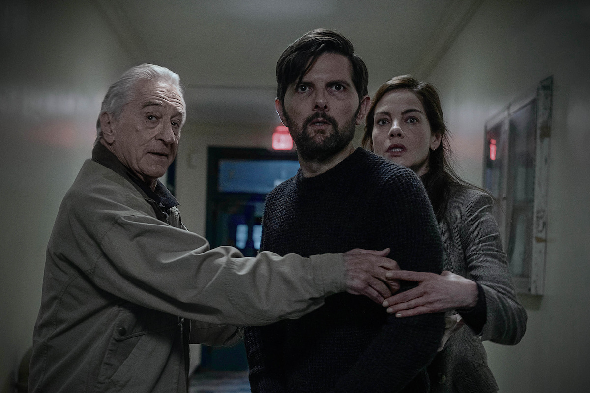 Three people standing close together in a dimly lit hallway, looking concerned or startled, with one man protectively reaching toward the others.