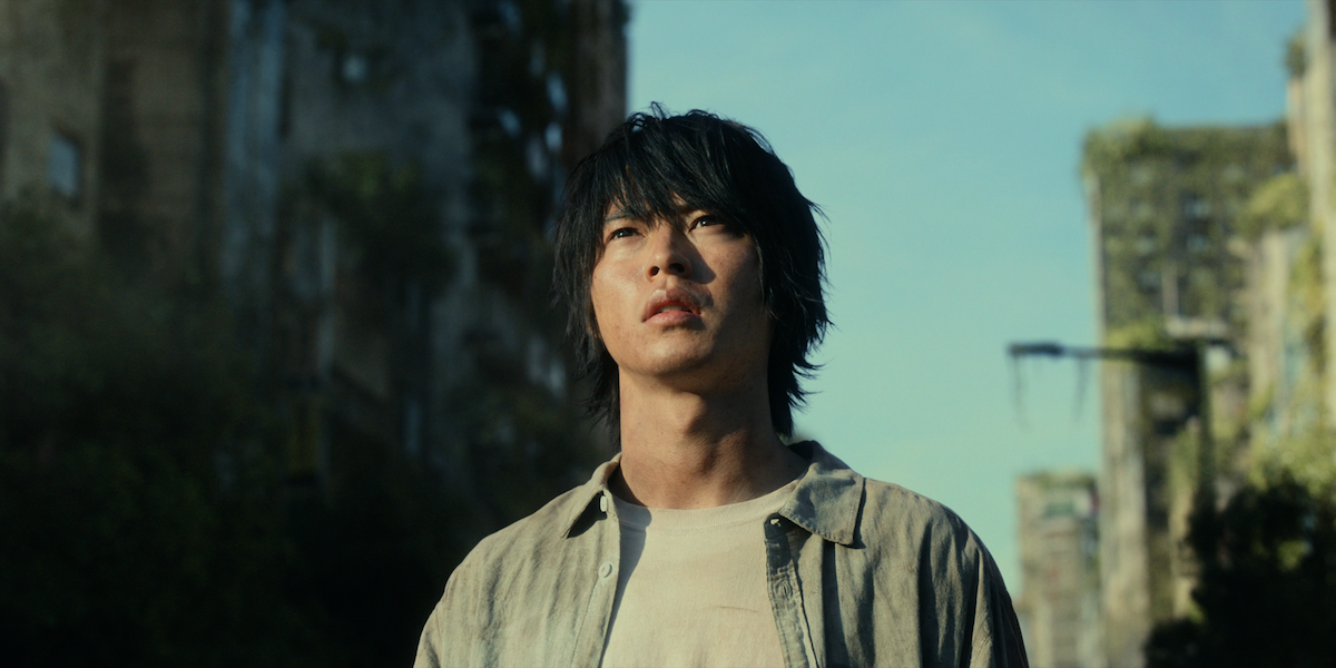 A young man with messy dark hair stands outside, looking up in a deserted urban area with overgrown, crumbling buildings under a clear sky, suggesting a post-apocalyptic or abandoned city environment.