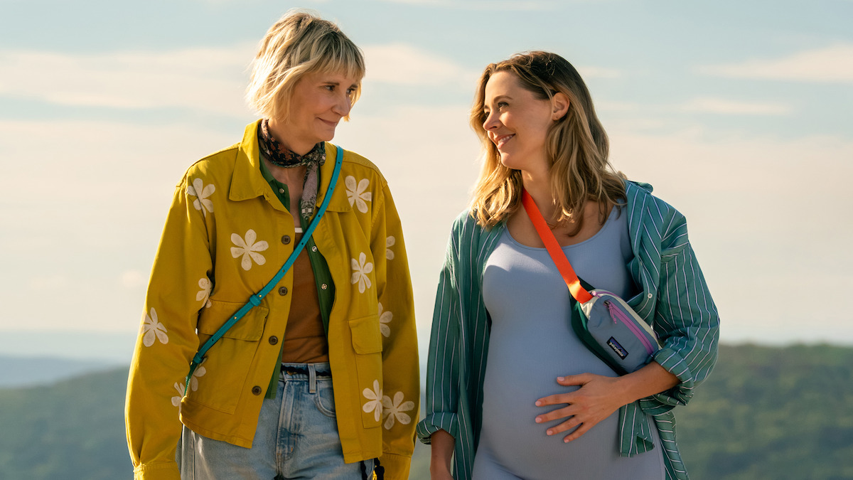 Two women walking outdoors on a sunny day, one visibly pregnant and smiling, both wearing casual clothes with light jackets, enjoying a scenic natural landscape in the background.