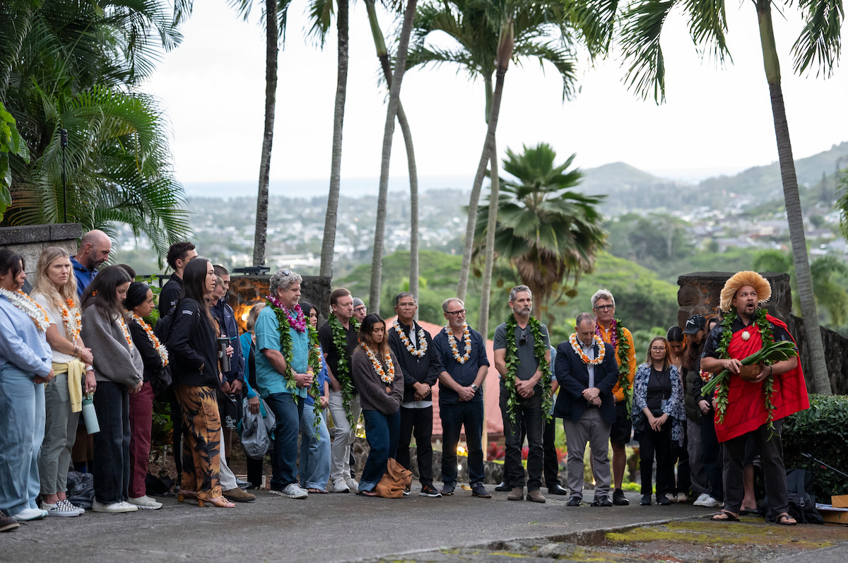 A group of people stands outdoors in a tropical setting with palm trees, listening to a person in traditional clothing holding a conch shell, with mountains and cityscape in the background.