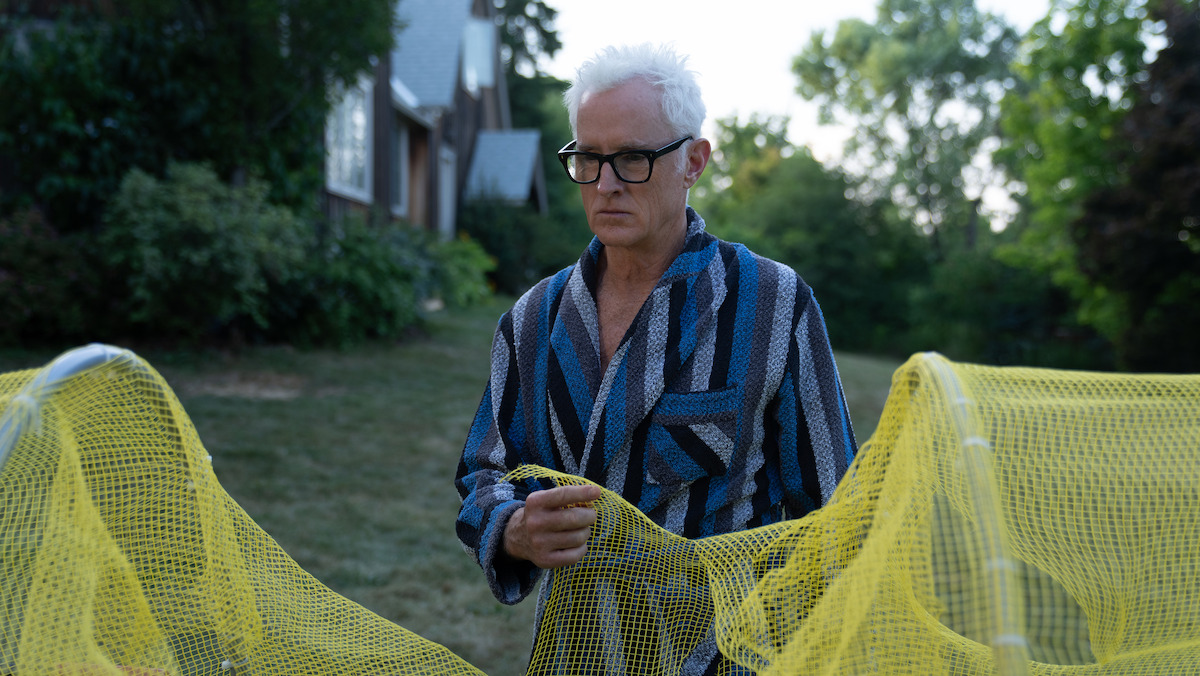 Older man with white hair and glasses in a blue striped robe stands outside on grass near house, handling yellow netting or mesh fencing, surrounded by trees and greenery in a residential backyard setting.