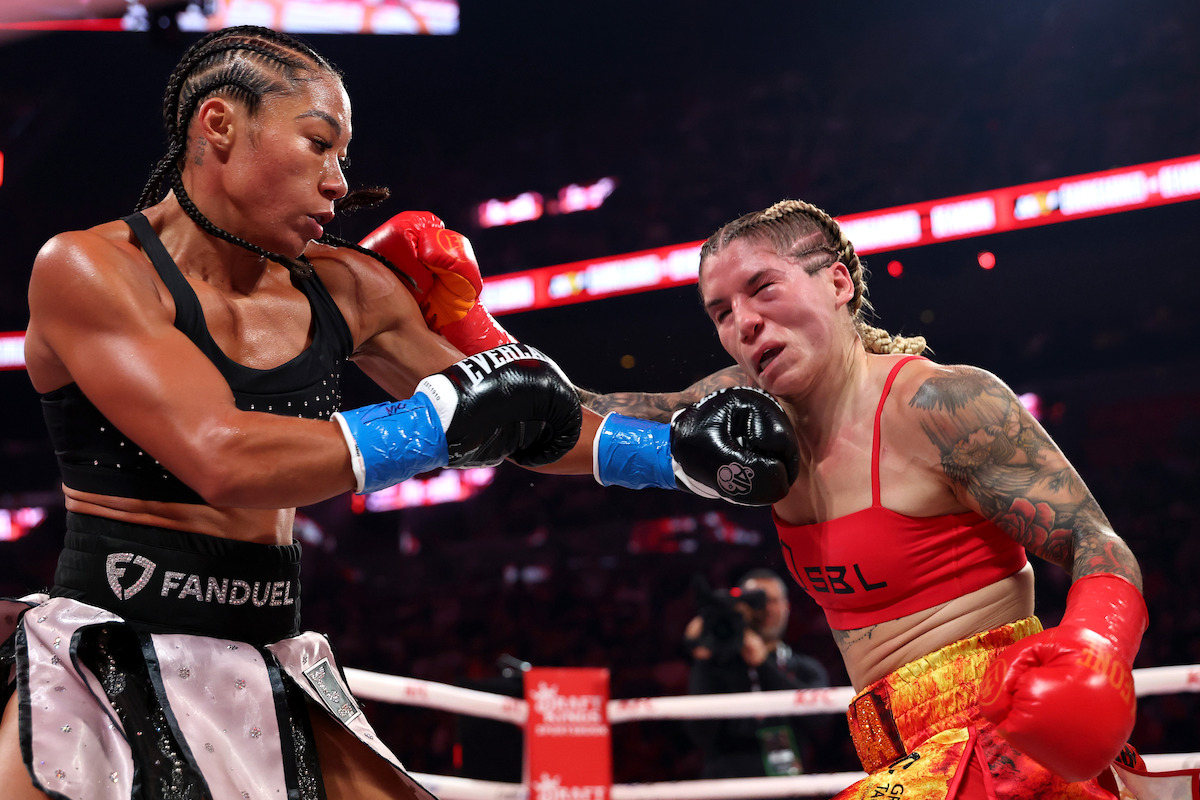 Alycia Baumgardner and Leila Beaudoin fighting in a brightly lit boxing ring, wearing gloves and sports gear, with a referee observing. The arena is filled with spectators and overhead lights.