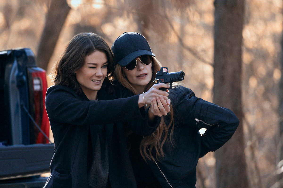 Two women in dark clothing stand outdoors in a wooded area; one is teaching or assisting the other in aiming a handgun. A pickup truck bed is visible behind them, and the background features blurred trees in daylight.
