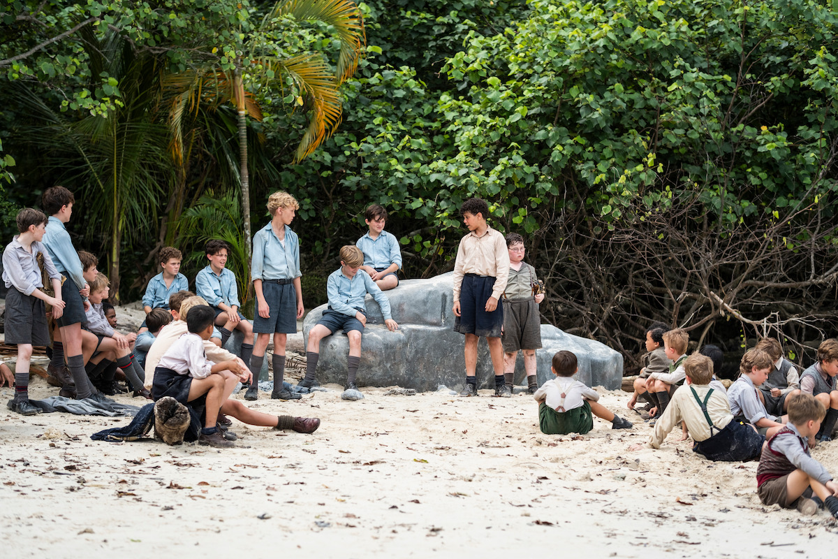 A group of boys in school uniforms gather on a sandy beach surrounded by lush green trees, some sitting on a large rock and others on the ground, appearing to be in a serious discussion or meeting in a tropical outdoor setting.