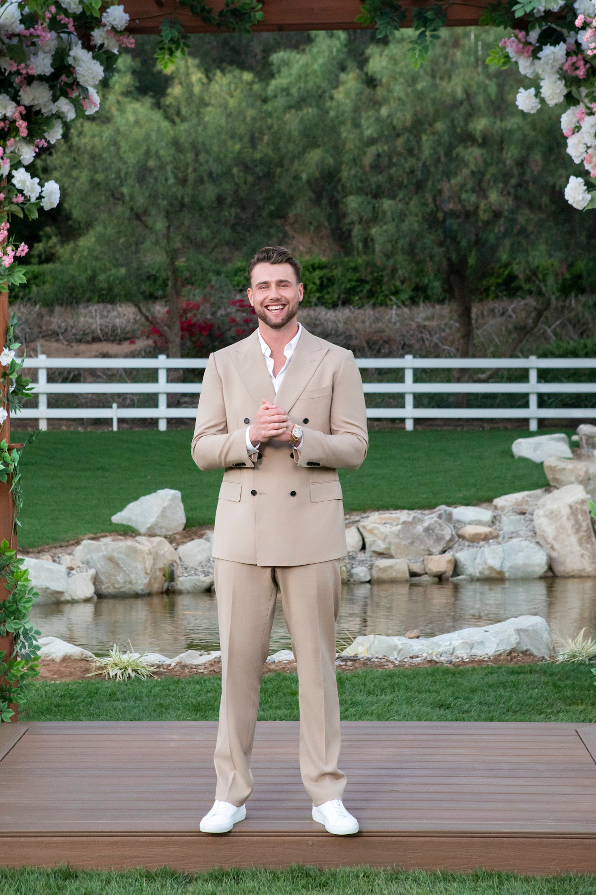 A smiling man in a beige suit stands on a wooden platform outdoors, framed by floral decorations, with a pond, rocks, green grass, white fence, and trees in the background on a sunny day.