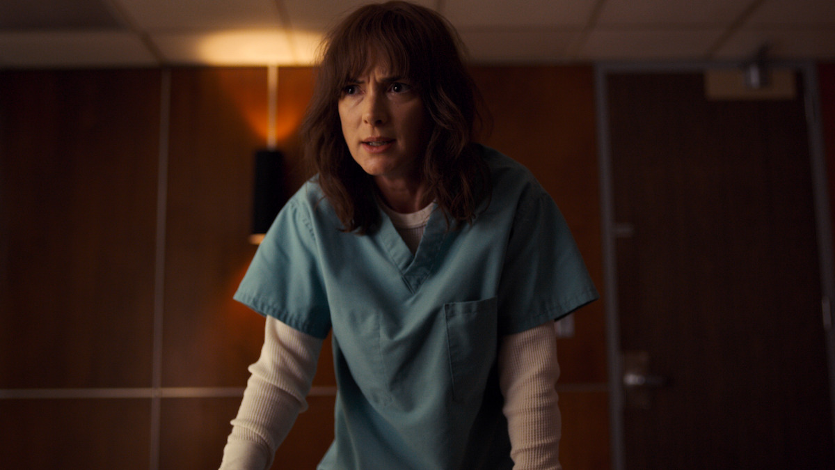 A woman in medical scrubs stands in a dimly lit room, leaning forward with a serious and intense expression, wooden wall panels and a closed door in the background, creating a tense atmosphere.