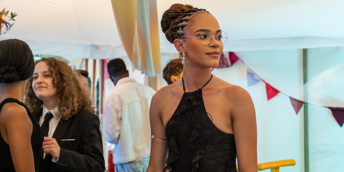 A woman in a black halter dress with glasses stands at a festive indoor event with other guests, bunting decorations, and a lively atmosphere in the background.