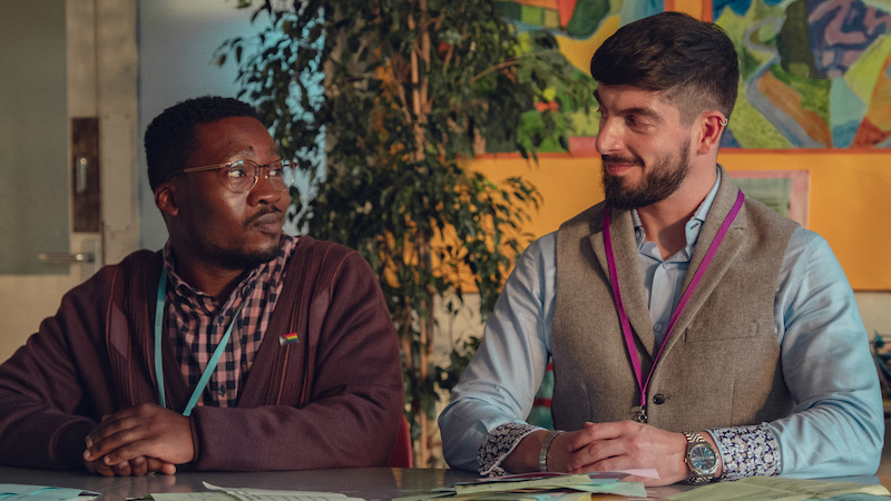 Two men sit at a desk in a colorful classroom setting, facing each other and smiling, with papers on the desk and artwork and a large plant in the background.