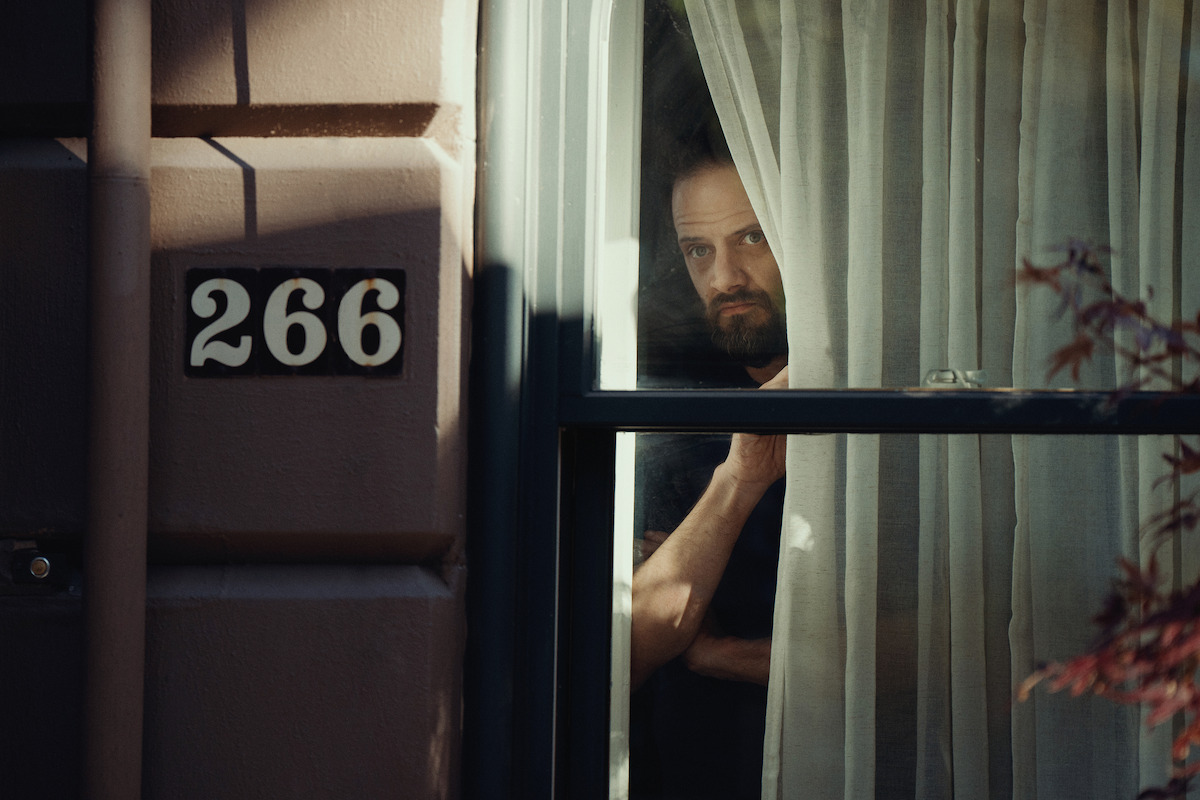 A man with a serious expression peers through sheer curtains from a window of a building numbered 266, with soft natural light and a moody, mysterious atmosphere.