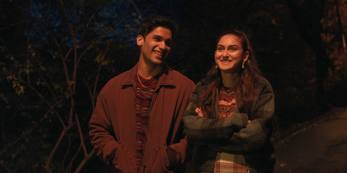 Two young adults walking together at night on a tree-lined path, both smiling and wearing warm jackets and sweaters, illuminated by streetlights in a relaxed outdoor setting.