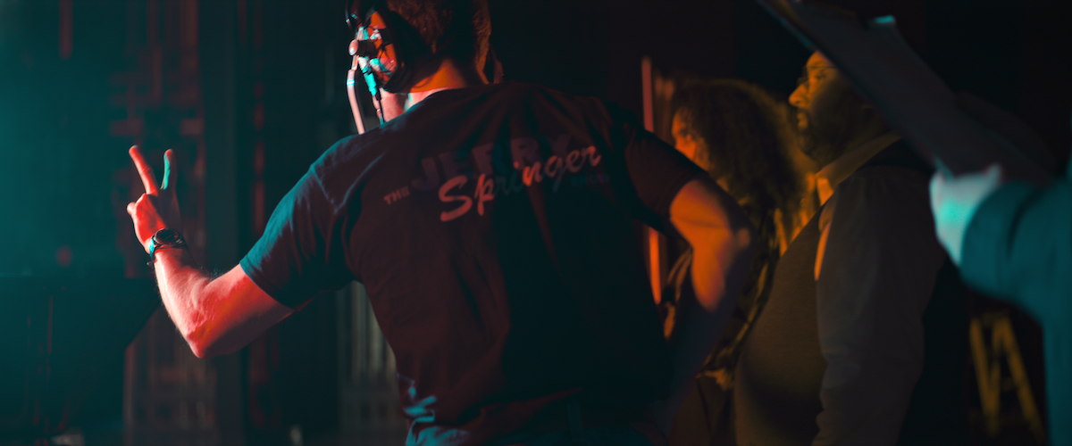 A man with a Jerry Springer Show t-shirt holds up two fingers backstage.