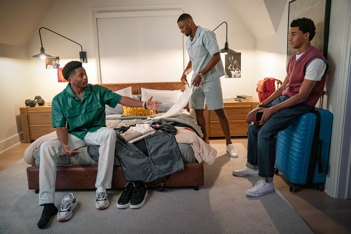 Three men in a modern bedroom, one sitting on the bed with clothes, another folding laundry, and the third sitting on a blue suitcase by the wall. Warm lighting and casual atmosphere suggest packing or preparing for a trip.