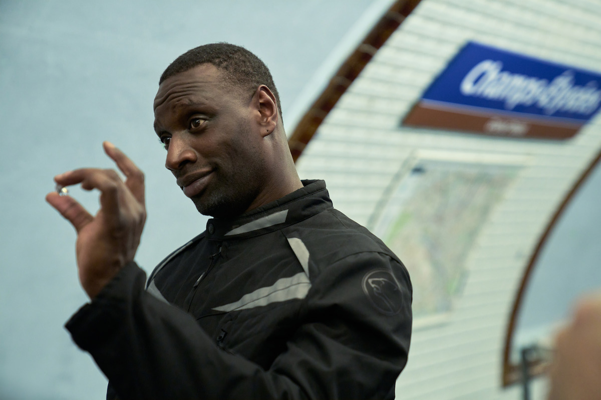 Man in black jacket making a hand gesture in a subway station, with a blue and white station sign and tiled wall in the background.