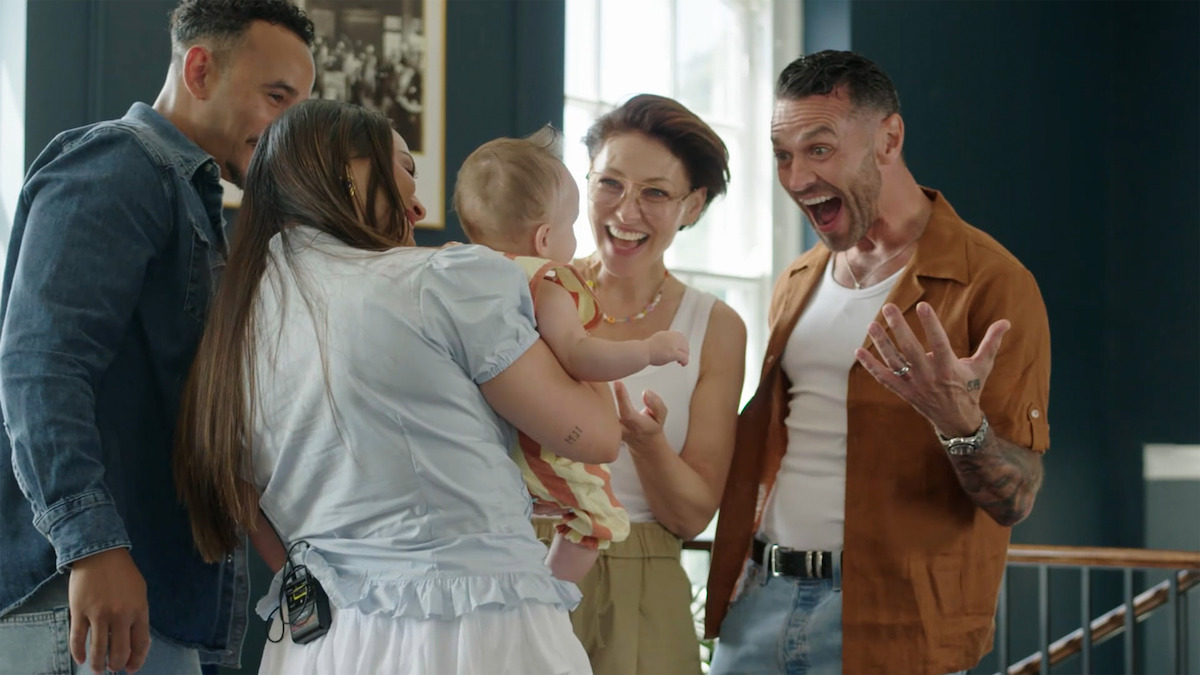 Four adults gathered indoors around a smiling baby, sharing a joyful moment in a brightly lit room with dark walls and framed pictures, one person expressing surprise and excitement.