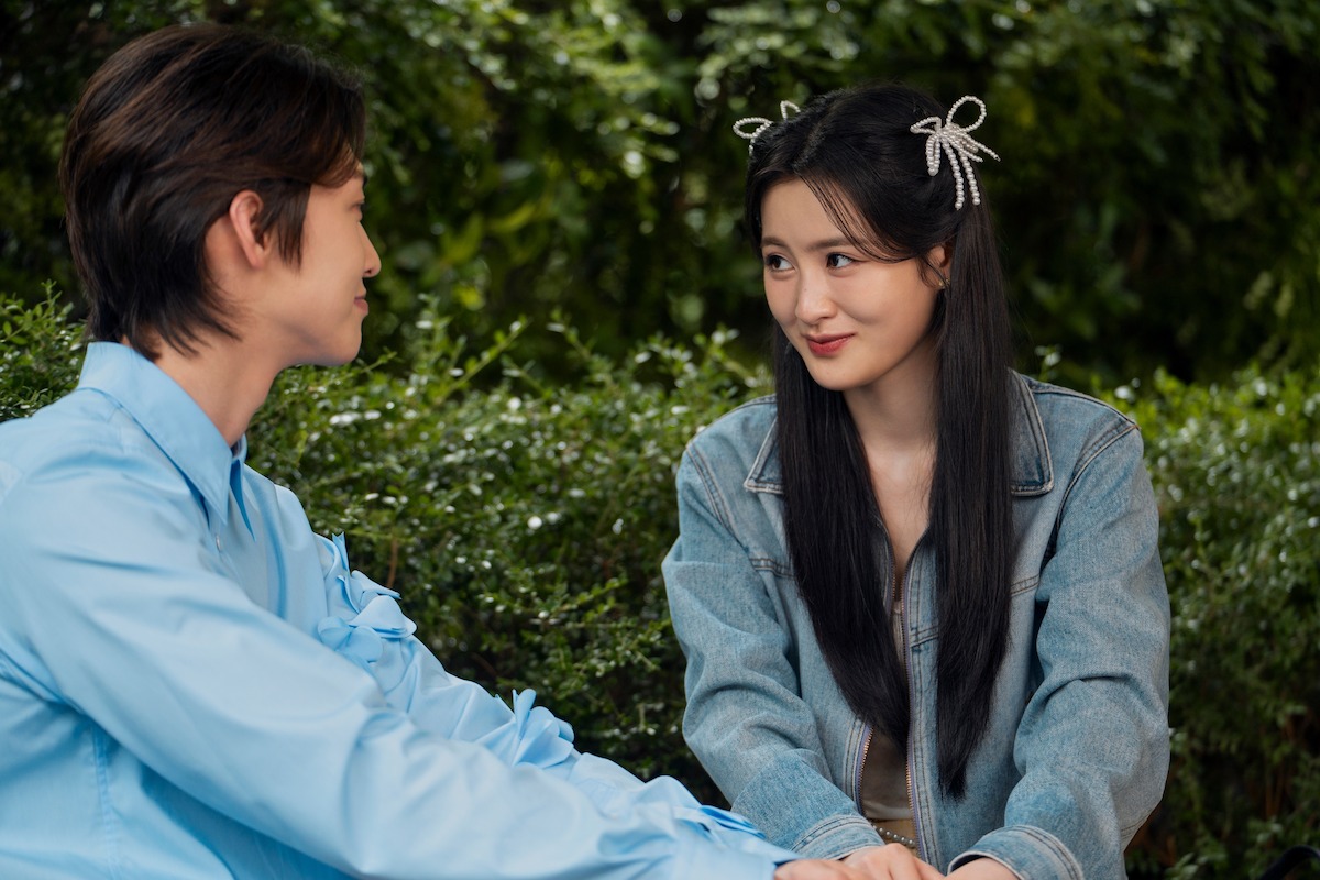Two young adults sit outdoors on a bench, surrounded by green bushes. They face each other, smiling and holding hands, with a relaxed and friendly atmosphere.