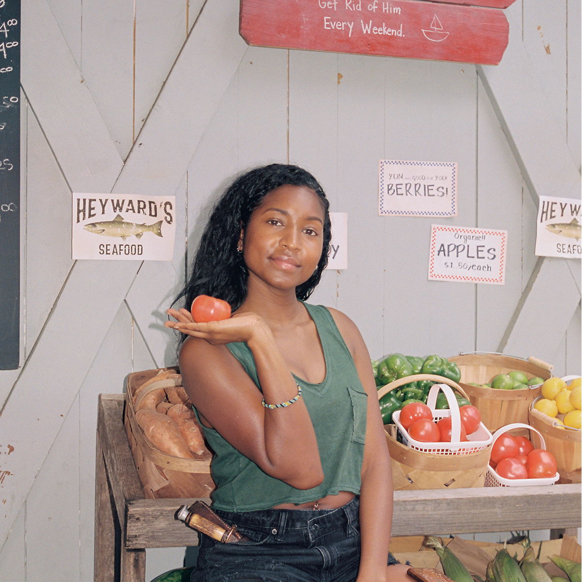 Woman in a green tank top at a farmer's market stand holding a tomato, with baskets of vegetables around her and signs for seafood, berries, and apples on a wooden background.