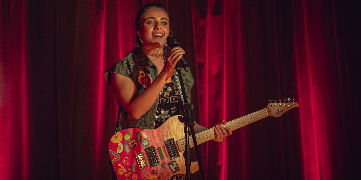 Smiling young woman stands on stage in front of red curtains, holding a pink electric guitar decorated with stickers and singing or speaking into a microphone during a live performance or talent show.