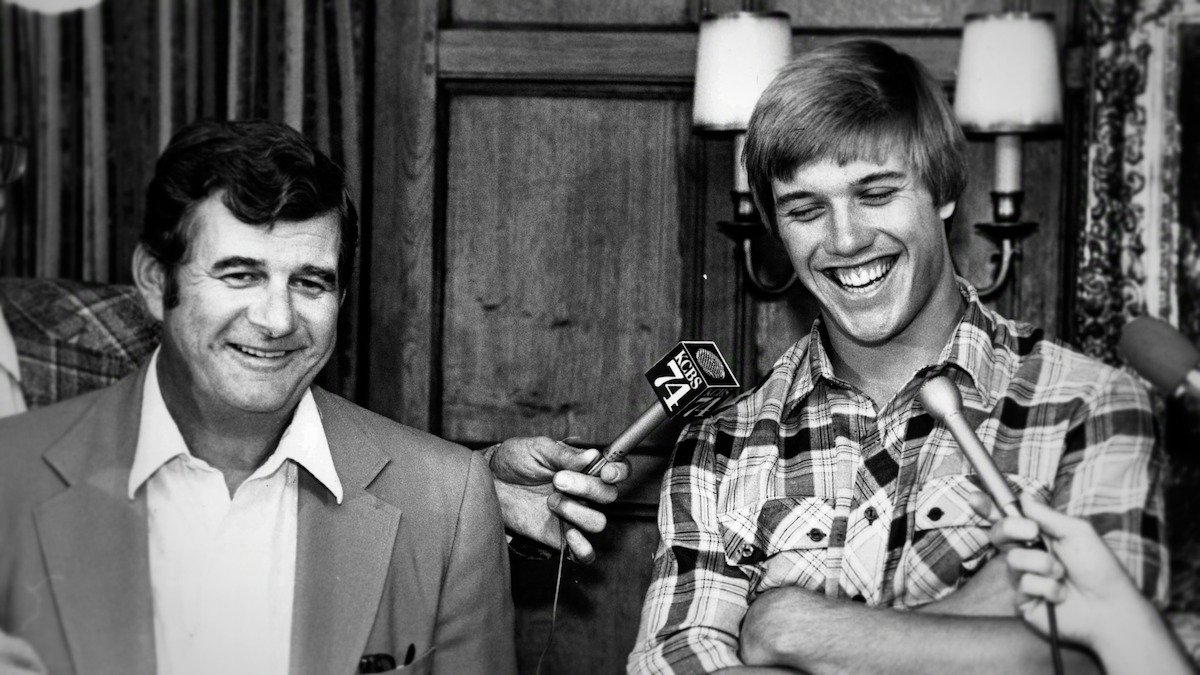 Jack and John Elway smiling during a press interview; the younger man laughs while holding his arms, with microphones pointed at both in a warmly lit indoor setting with wood paneling and lamps in the background.
