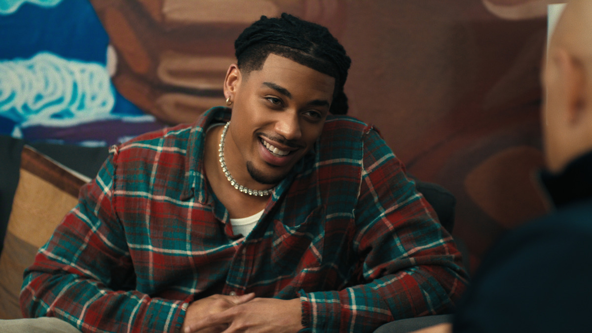 Smiling man in a plaid shirt sitting indoors, leaning forward in conversation with another person, colorful mural in the background.