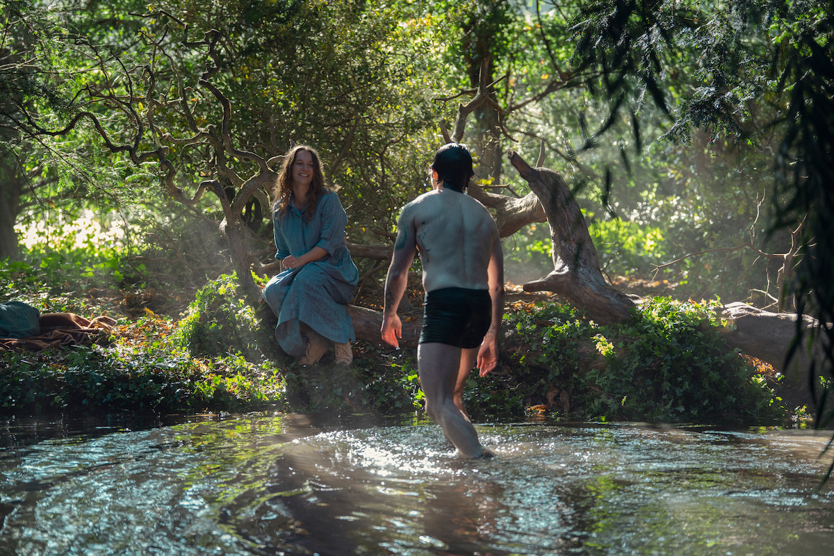 A woman sits on a tree while a man stands in a sunlit forest pond. Surrounding them are lush green trees and dappled light, creating a peaceful, natural setting.