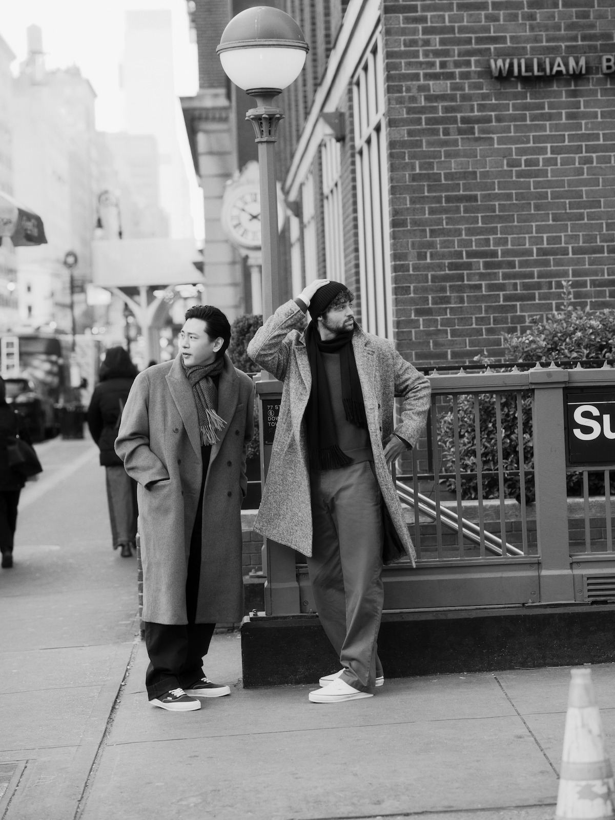 Teo Yoo and Noah Centineo stand on the street outside a subway entrance.