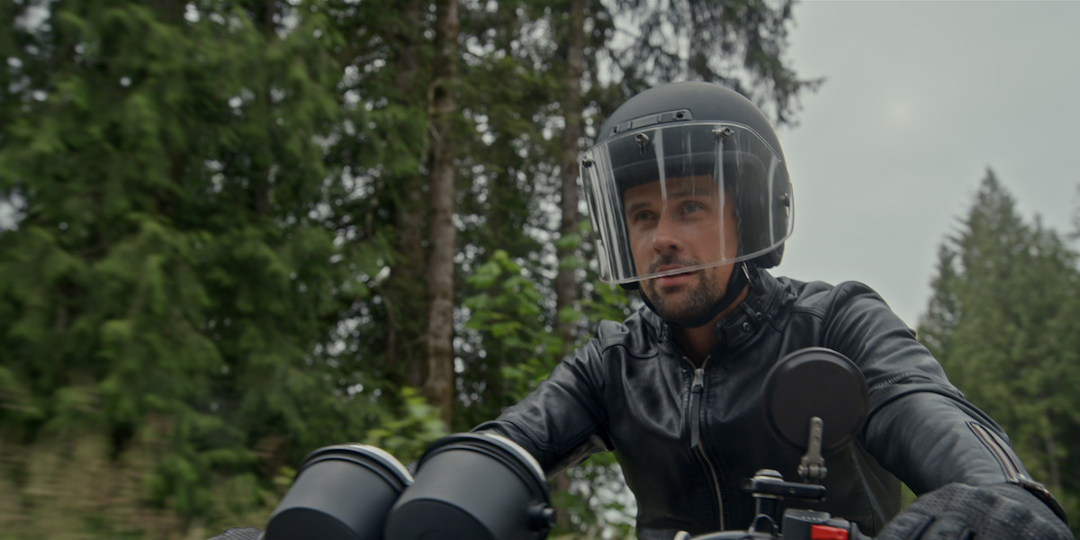 Man wearing a helmet and leather jacket riding a motorcycle through a forested area, surrounded by tall green trees under a cloudy sky.