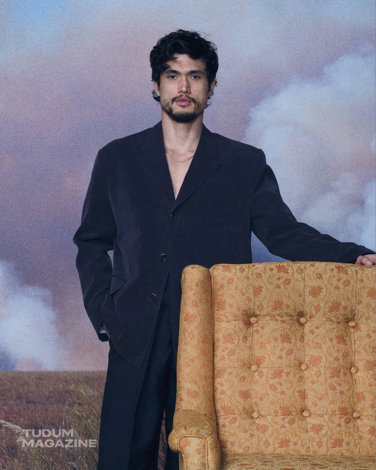 A man with dark hair and a beard wearing a navy blazer and gold chains stands in front of a blurred outdoor landscape with cloudy sky, and a vintage floral pattern armchair is next to him.