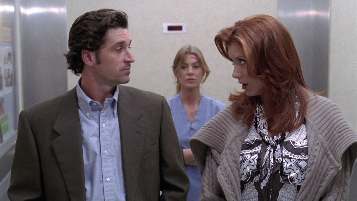Man in a suit and woman in patterned outfit talking in a hospital hallway, with a woman in scrubs and a bandage on her forehead standing behind them. Neutral colors and medical setting.