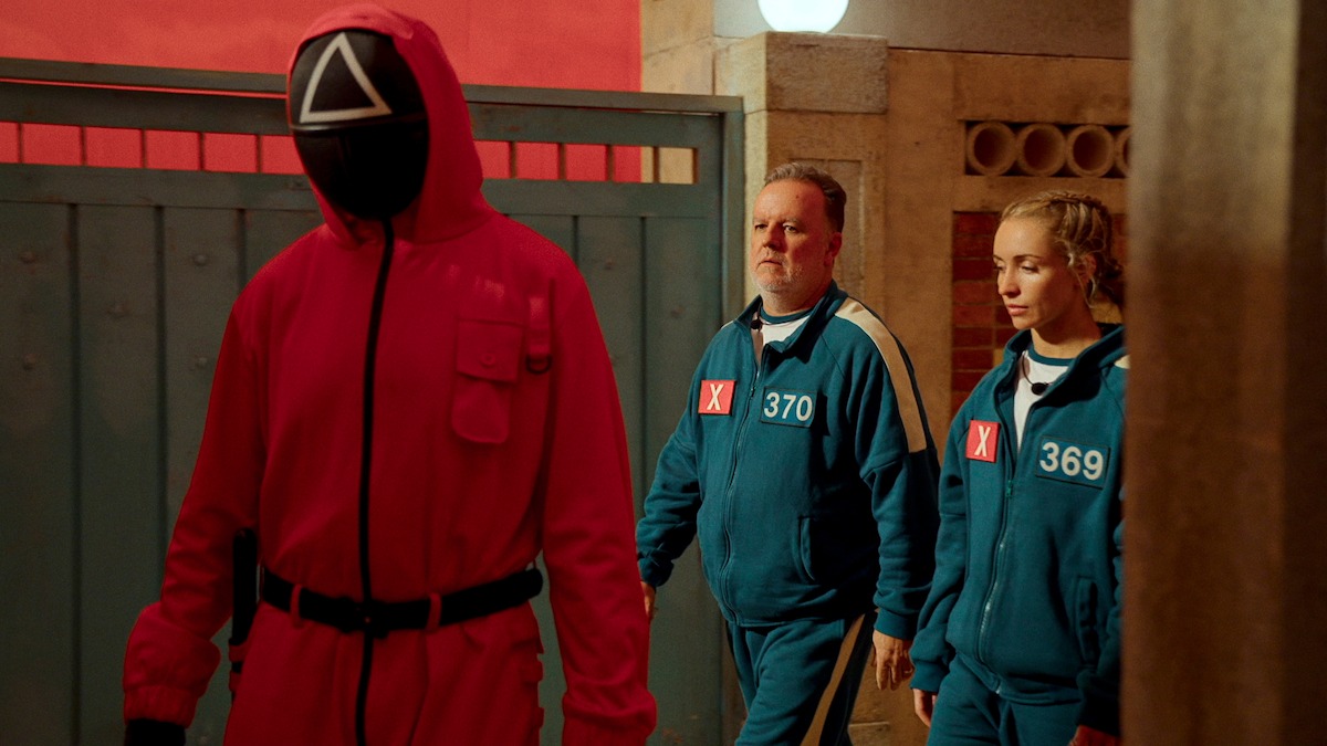 A guard in a red jumpsuit and mask leads two contestants in blue tracksuits with numbers through a brightly lit corridor, creating a tense and mysterious atmosphere.