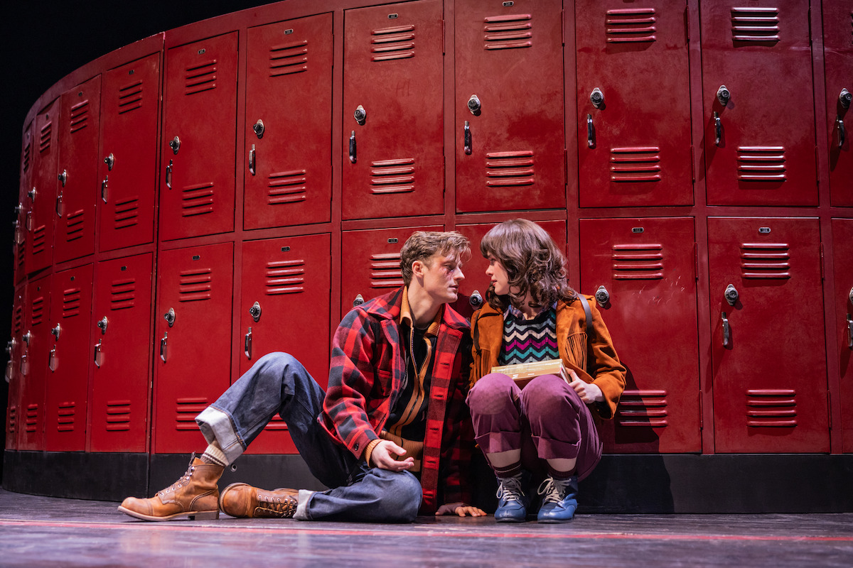 Two people sit on the floor in front of a row of red lockers, dressed in casual clothes, looking at each other closely on a stage, suggesting a dramatic or emotional scene set in a school environment.