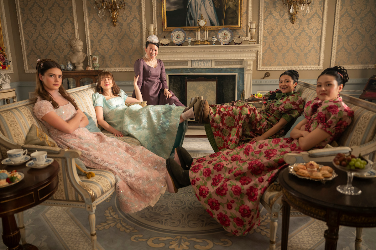 Năm người phụ nữ trong những bộ váy thanh lịch thời xưa ngồi trong một phòng khách được trang trí lộng lẫy với giấy dán tường cầu kỳ, đồ nội thất cổ điển và bộ ấm trà, thư giãn với đôi chân gác lên bàn, gợi lên một buổi gặp gỡ xã giao thoải mái và vui vẻ.