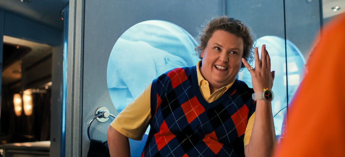 Smiling person in a colorful argyle vest and yellow shirt stands in front of a blue-lit background with circular windows, appearing to be inside a modern building or transportation interior.