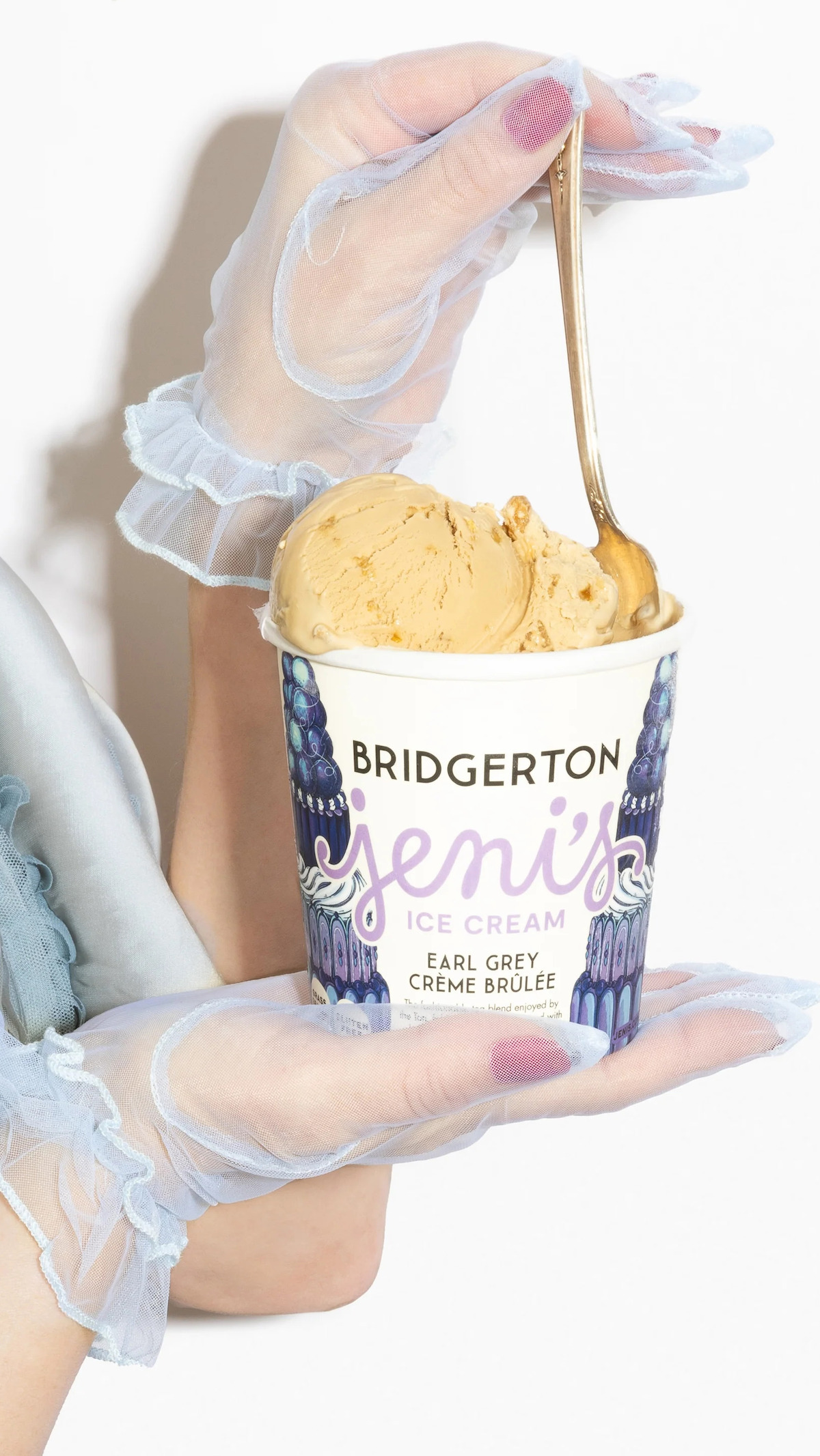 A hand in sheer, frilly gloves holds a cup of Jeni’s Bridgerton Earl Grey Crème Brûlée ice cream, with a spoon scooping ice cream, against a bright, minimal background.