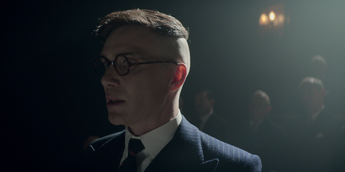 A man in 1920's attire and glasses stands up in a crowded hall which is dimly lit.