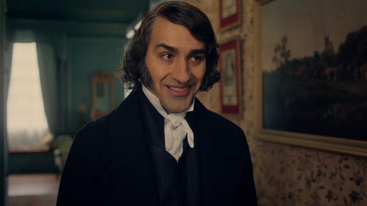 A man in early 19th-century attire with sideburns smiles in a warmly lit, vintage room with floral wallpaper, framed paintings, and a window in the background.