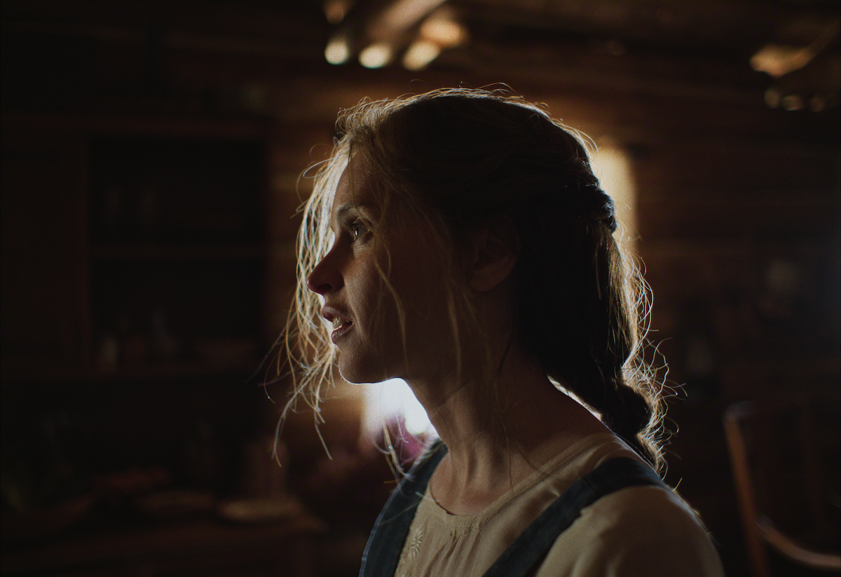 A young woman with messy hair stands in soft light inside a rustic wooden room, gazing thoughtfully into the distance. The environment feels quiet and reflective, with warm, dim lighting enhancing a nostalgic mood.
