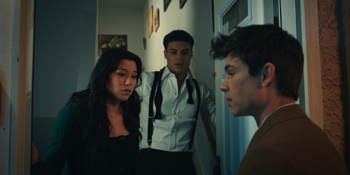 Three young adults standing in a hallway near a door, one woman and two men, appearing concerned and engaged in a serious conversation, indoors with dim lighting and paintings on the walls.