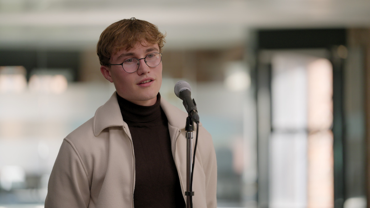 Young man with glasses and light brown hair stands indoors in front of a microphone, wearing a cream jacket and dark turtleneck, in a softly lit modern setting with a calm, focused mood.