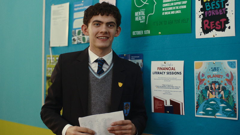 A student in a school uniform stands indoors holding papers, smiling in front of a blue bulletin board with posters about mental health, financial literacy, and environmental awareness.