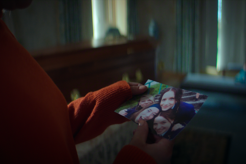 Person in an orange sweater holds a photo of three smiling women in an indoor room with a closed wooden casket in the background, suggesting a memorial or funeral setting.