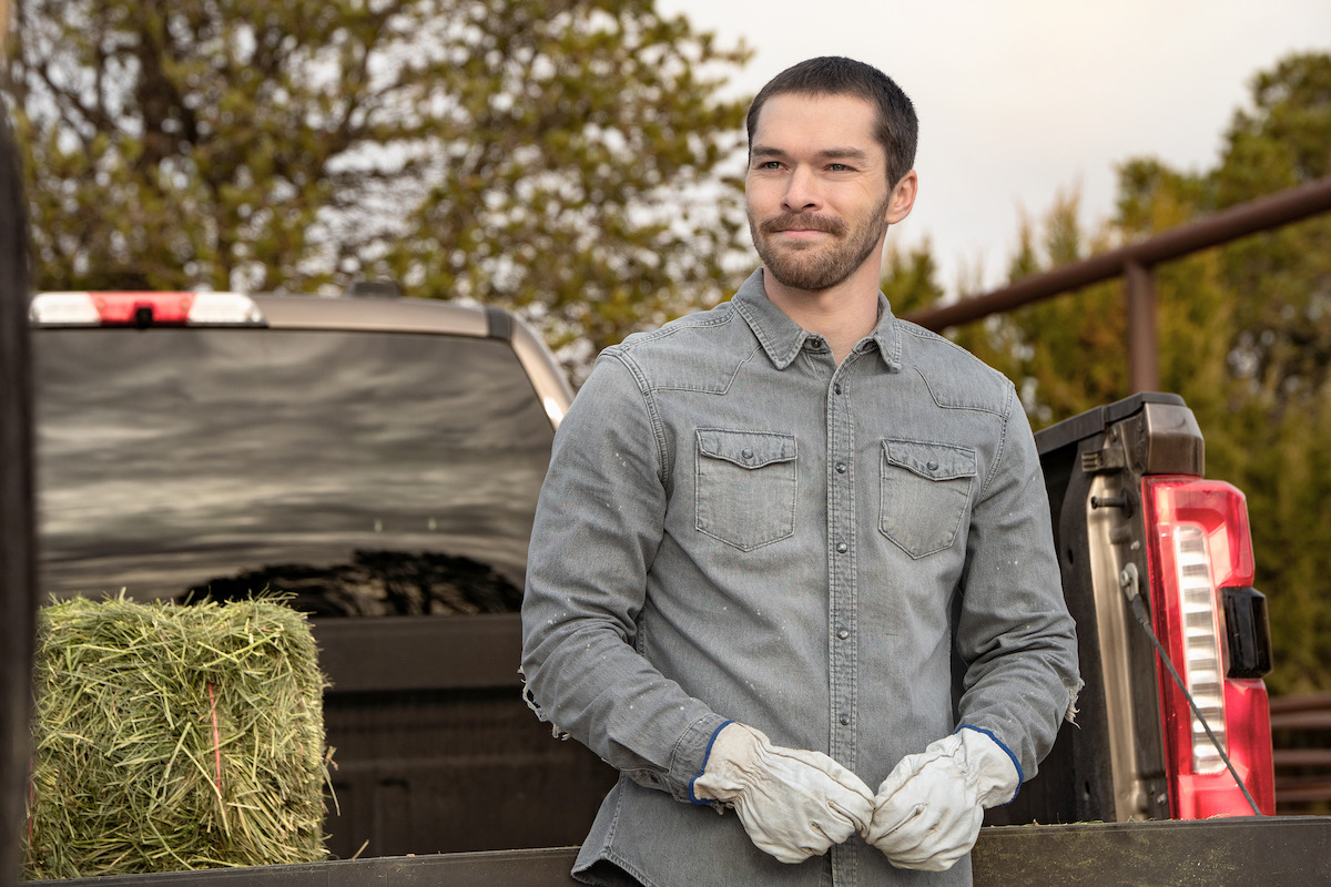 Man wearing work gloves and denim shirt stands next to a pickup truck with a hay bale in the truck bed, outdoors on a farm with trees and fencing in the background.