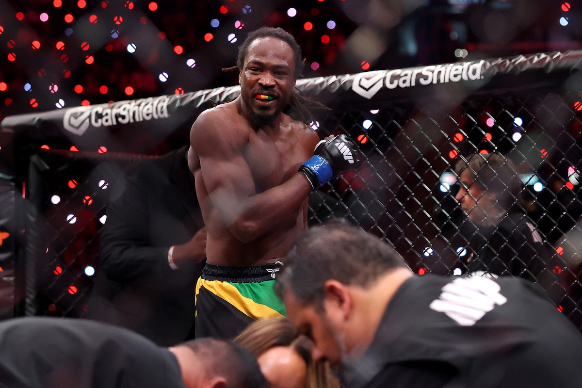 Mixed martial artist standing in an octagon cage, wearing gloves and shorts with a Jamaican flag design, while others attend to someone on the ground, under bright arena lights and a crowd in the background.