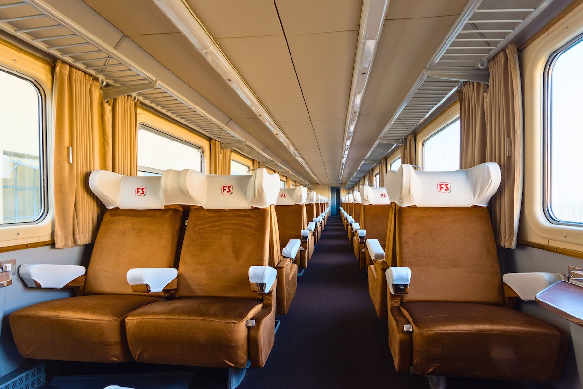 Empty vintage train carriage with brown cushioned seats, white headrests, wide windows with curtains, overhead luggage racks, and soft natural light creating a calm, comfortable, and inviting atmosphere.