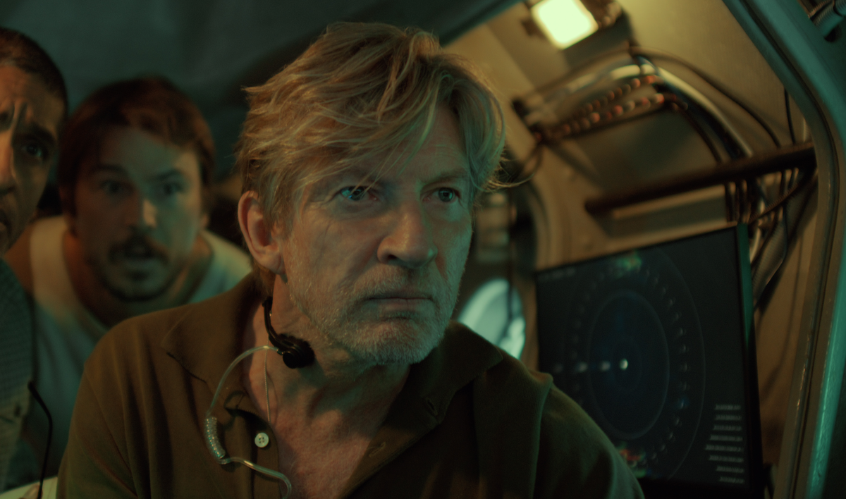 Three men inside a dimly lit submarine or control room, one wearing a headset, looking intently at a screen displaying radar or sonar imagery, with cables and equipment in the background.