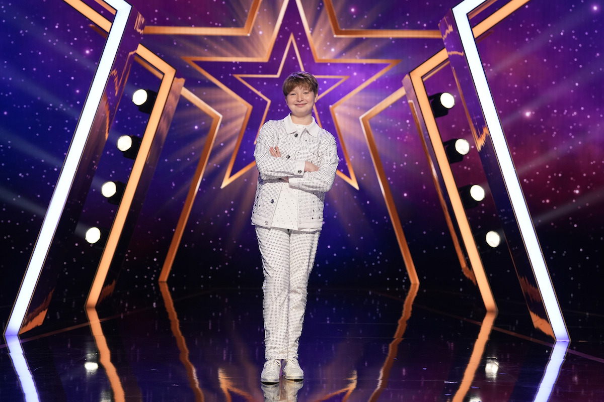 Yair Keydar stands confidently on a shiny stage with arms crossed, wearing a white outfit. The background features a large glowing star design with purple and gold colors and stage lights on either side.